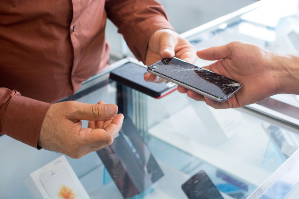 Person handing smartphone with smashed screen to other person above glass shop counter.